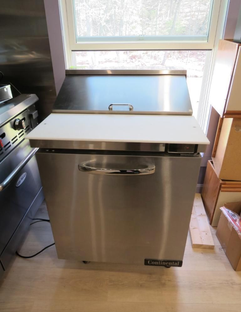 Stainless steel commercial sandwich prep refrigerator with open lid and cutting board in a kitchen.