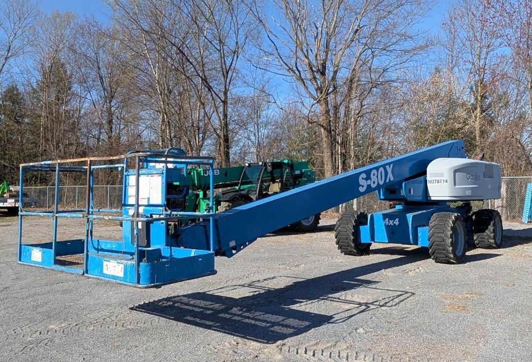 Blue boom lift parked on gravel beside leafless trees under a clear sky