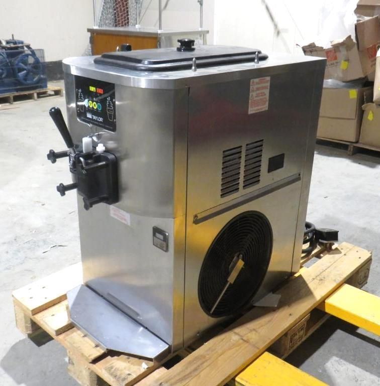 Stainless steel soft-serve ice cream machine on a wooden pallet. Black controls, dispensing handle, and cooling fan.