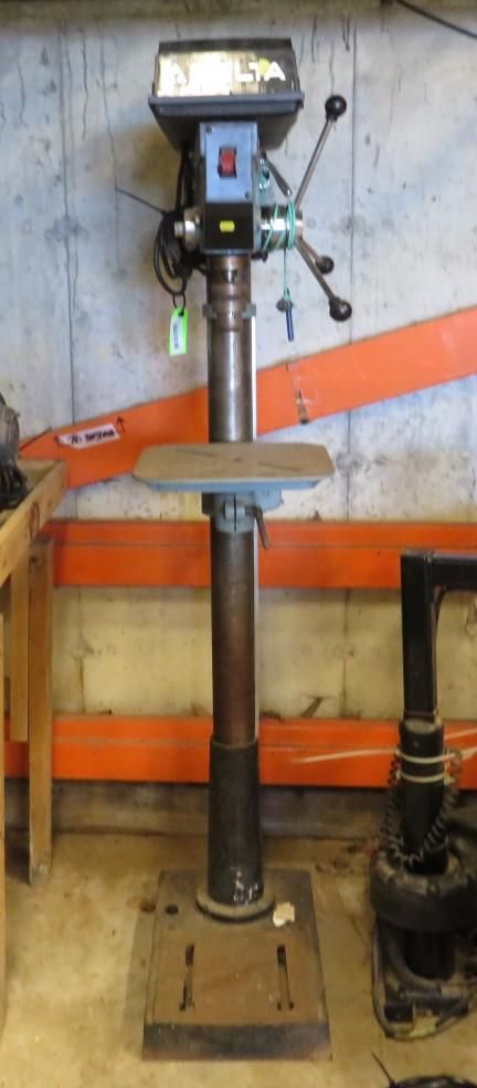 A floor-standing drill press with a metal base, column, and work table in a workshop setting.