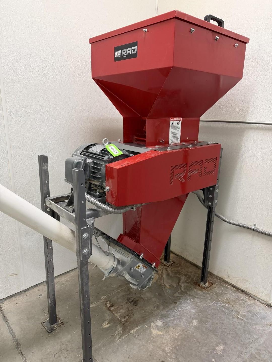 Red industrial grinder with hopper and auger, bolted to a frame, in a corner.