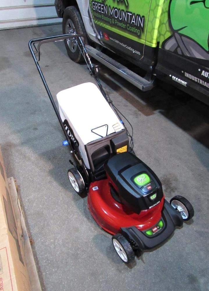 Red and black Toro lawnmower with a white battery and black handle, parked on concrete near a truck.