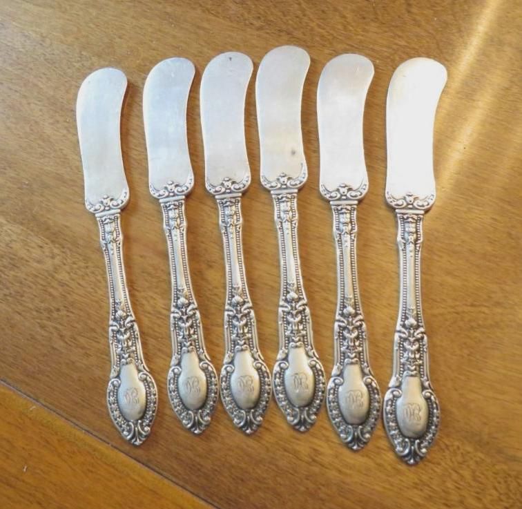 Six silver butter knives with ornate handles on a wooden table.