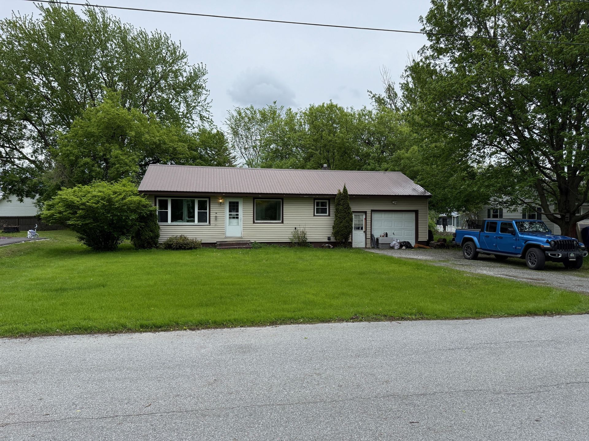 Beige house with a brown roof and attached garage, blue truck parked in driveway, overcast day.