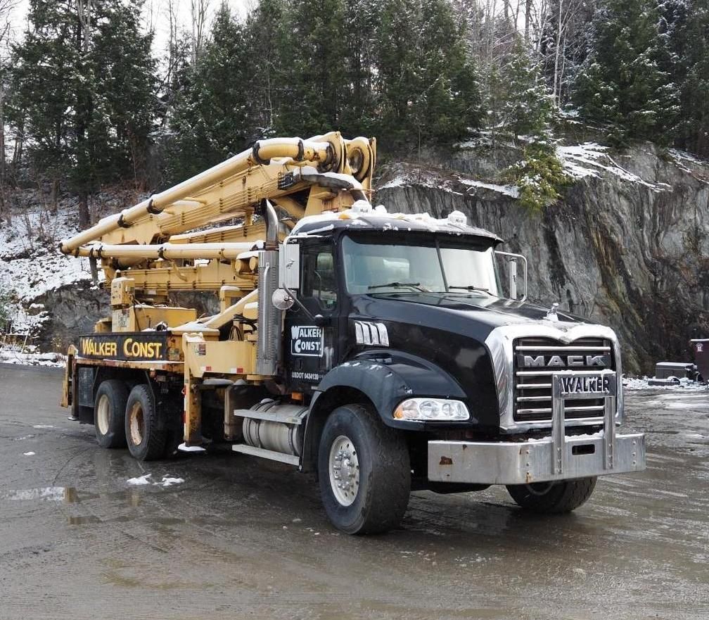 Black and yellow concrete pump truck parked on a snowy road with a forest and rock face in the background.