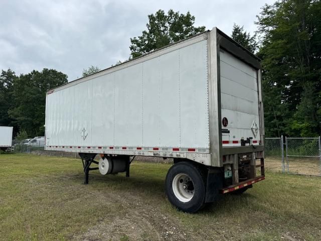 White semi-trailer parked on grassy ground with trees in background.