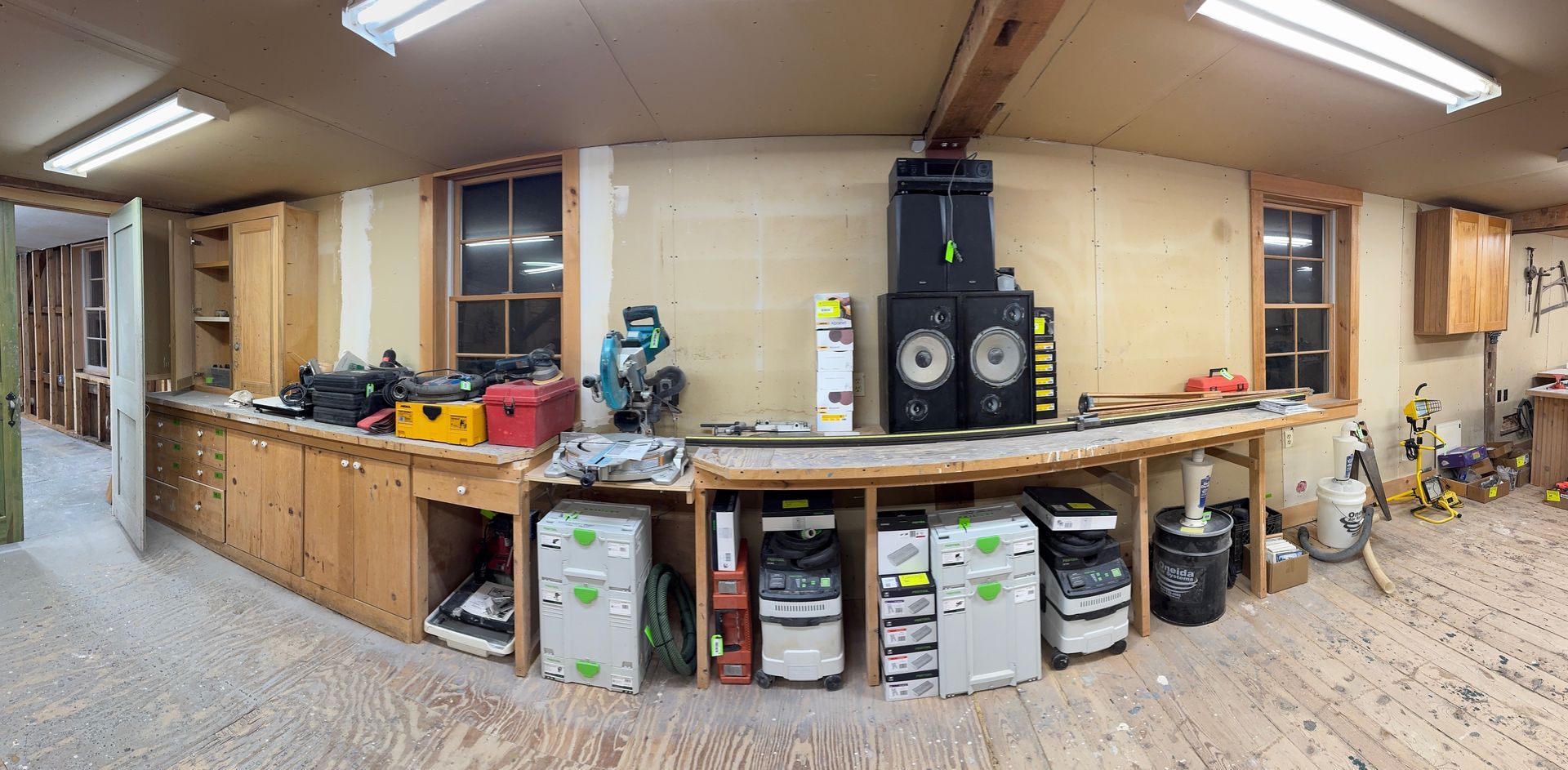 Wide workshop with long workbench, tools, storage shelves, and fluorescent lights throughout.