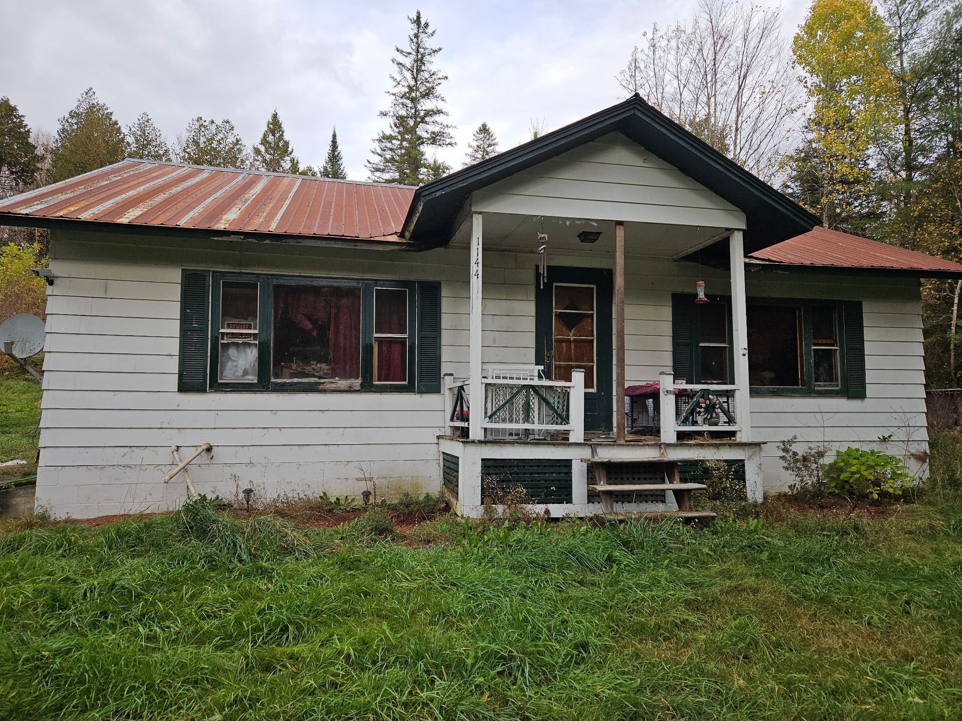 A one-story white house with a metal roof, dark shutters, and a small front porch in a wooded setting.