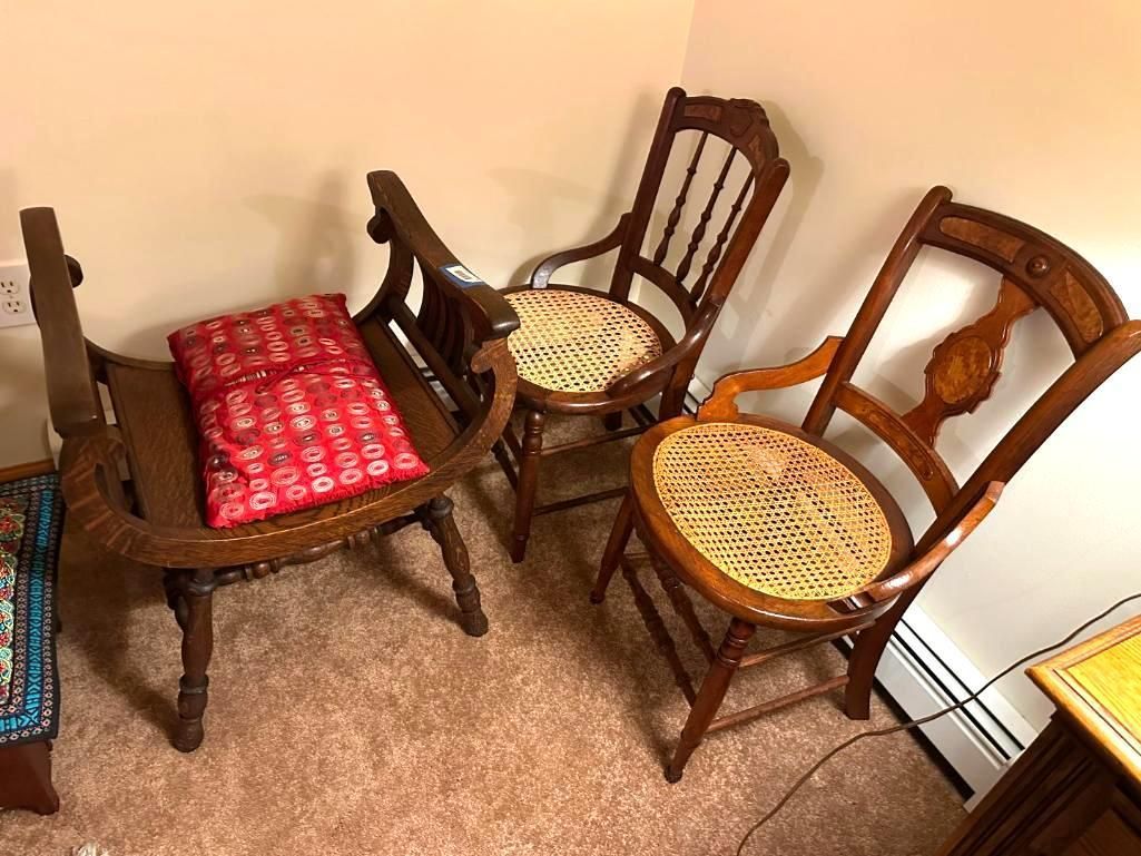 Three antique wooden chairs with woven seats, in a corner. One has a red cushion.