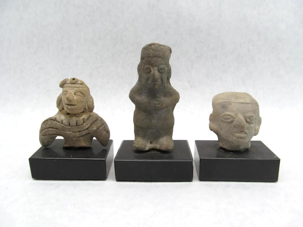 Three ancient clay figurines on black pedestals: a figure with wings, a standing figure, and a head.