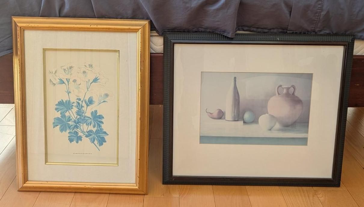 Two framed artworks on a couch: a blue floral print and a pastel still life with a vase and jug.
