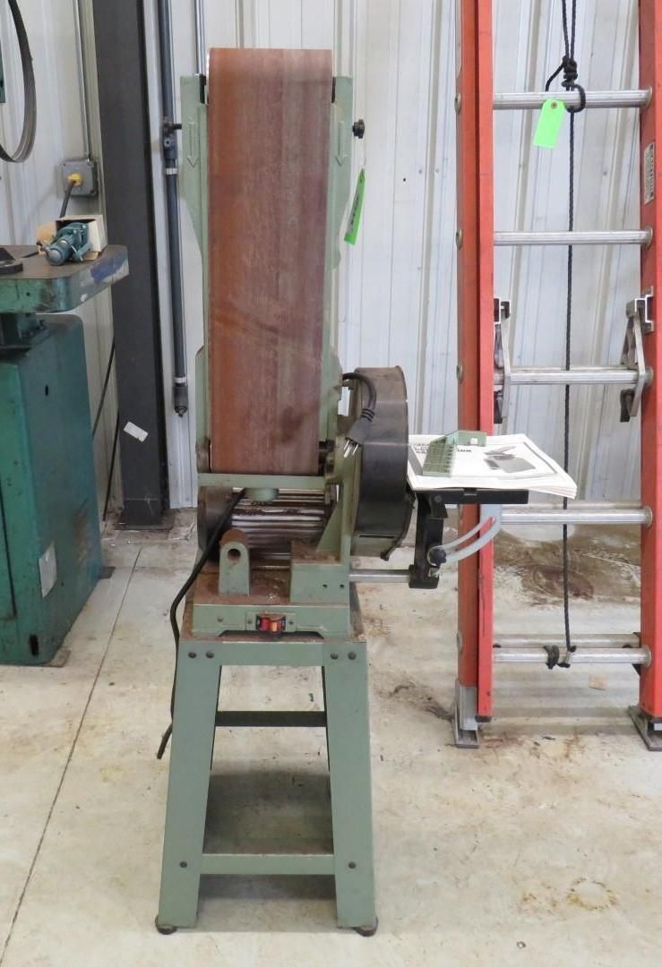 Green belt sander on a stand, against a wall with a ladder.