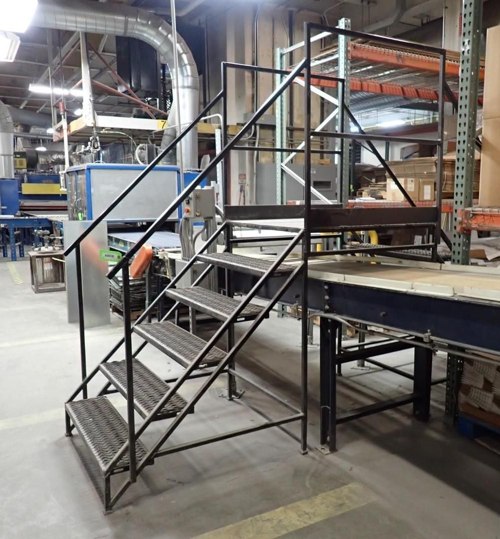 Metal staircase with railing and small platform in a factory setting, leading to a conveyor.