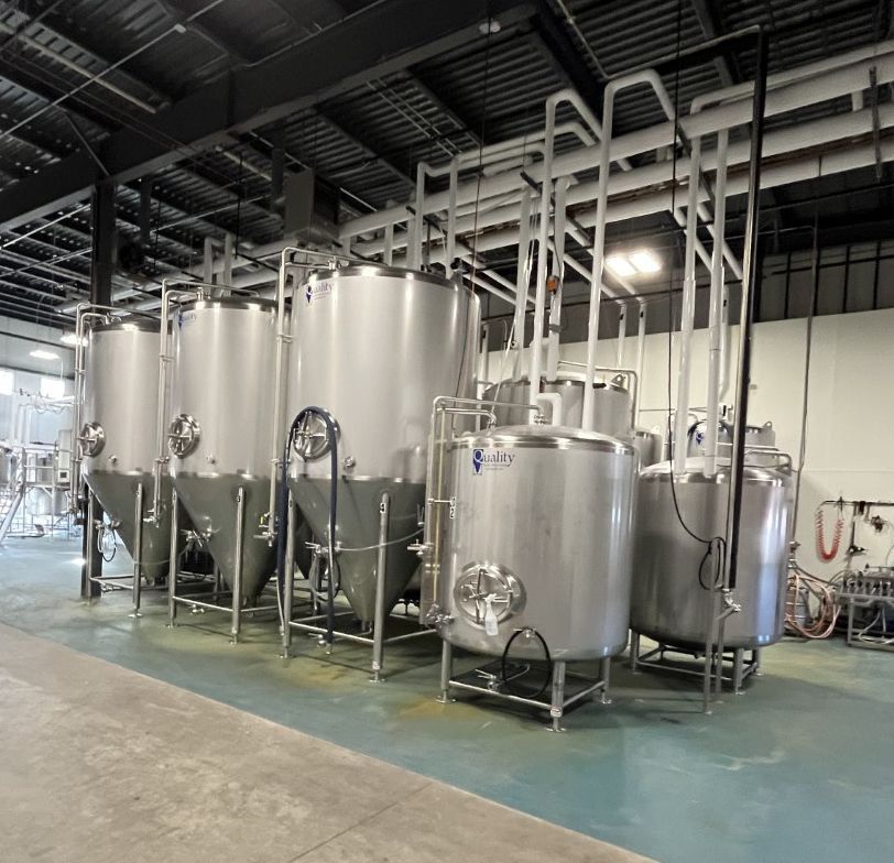 Stainless steel fermentation tanks in a brewery, with connecting pipes and a concrete floor.