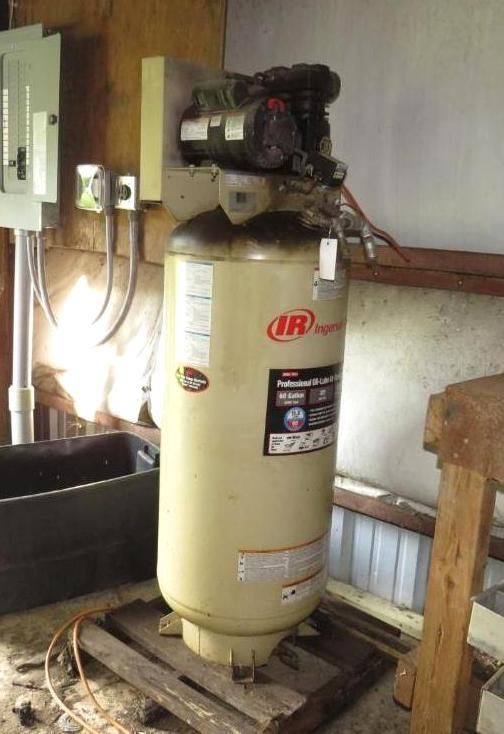 An air compressor in a shed, beige tank with black top, and electrical panel on wall.