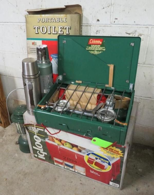 A collection of camping gear including a Coleman stove, Igloo cooler, portable toilet, and thermos bottles.