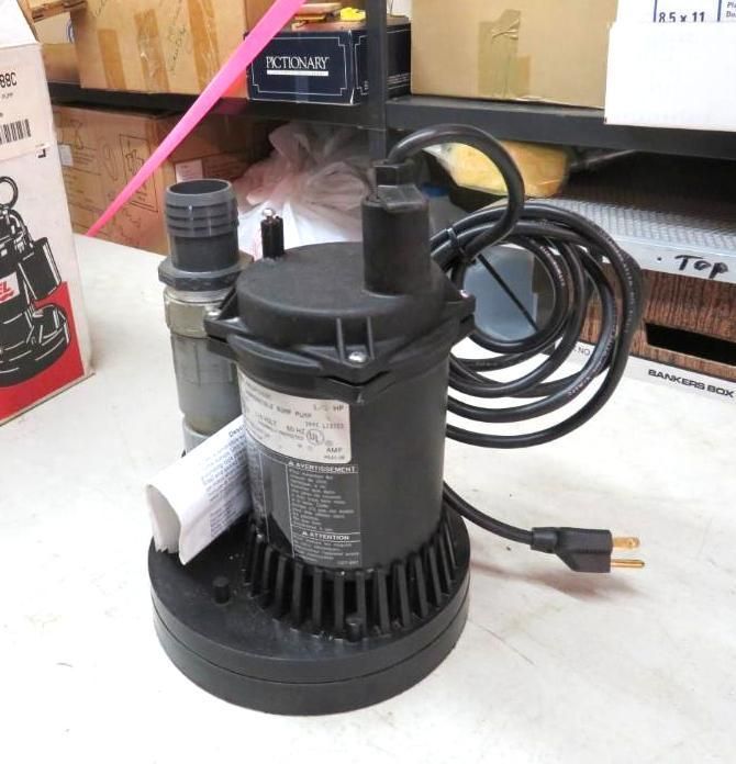 Black sump pump with power cord, attached to a pipe, on a white surface.