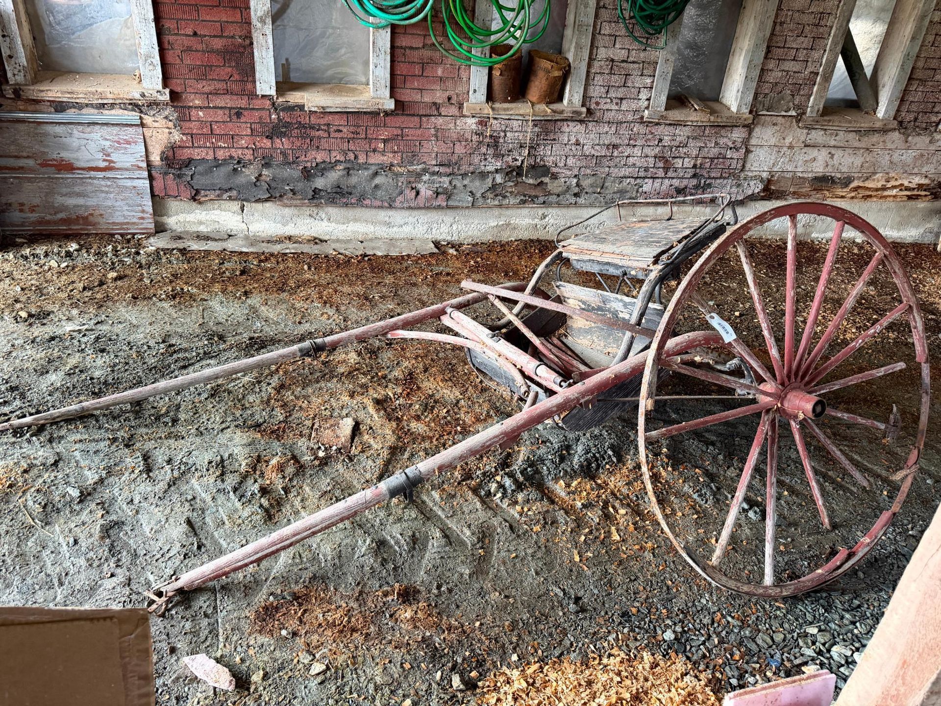 Old wooden handcart with a red wheel, lying on dirt floor beside a brick wall.