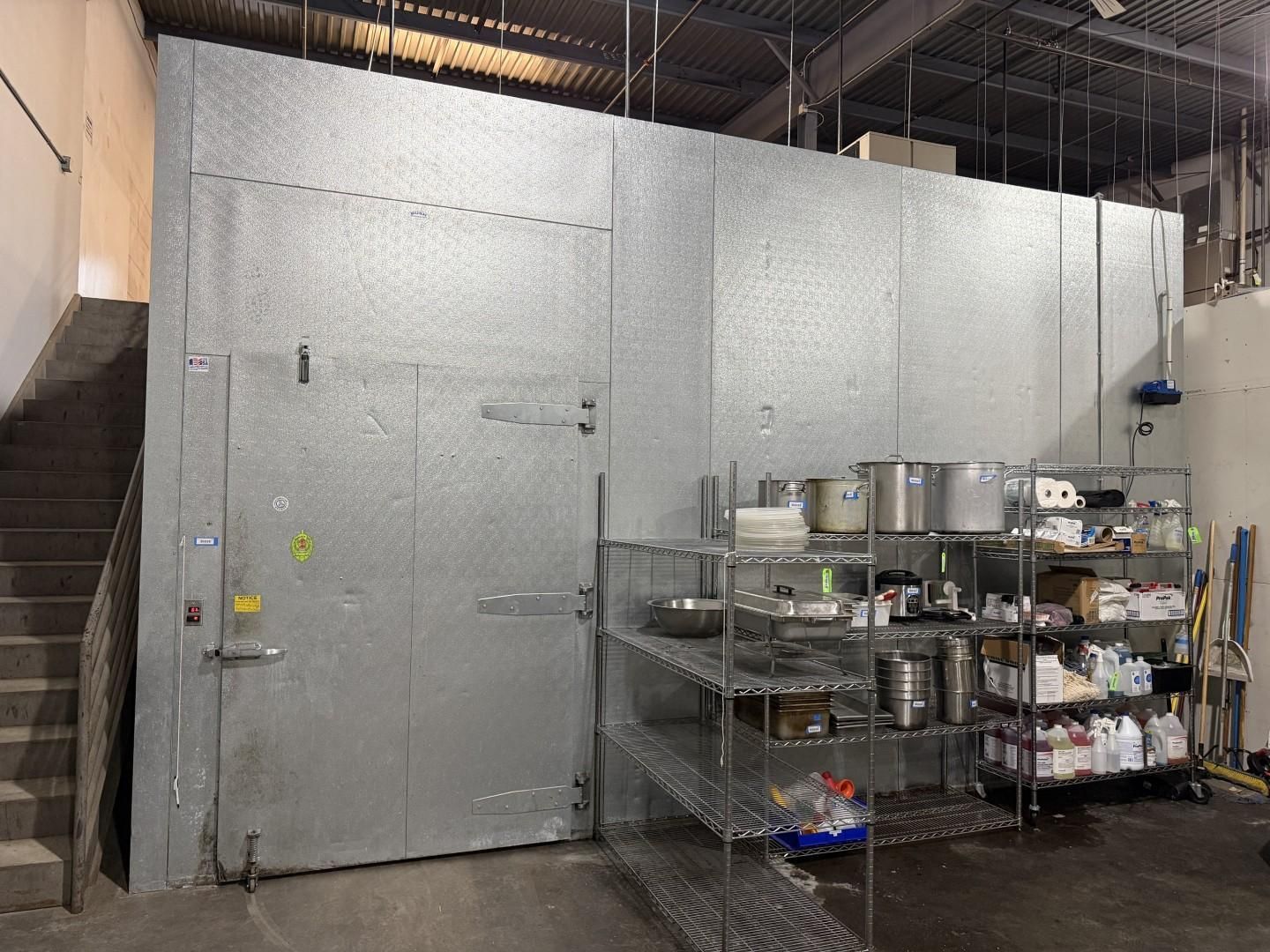 Large walk-in freezer with door and metal shelving containing kitchen supplies. Gray walls, industrial setting.