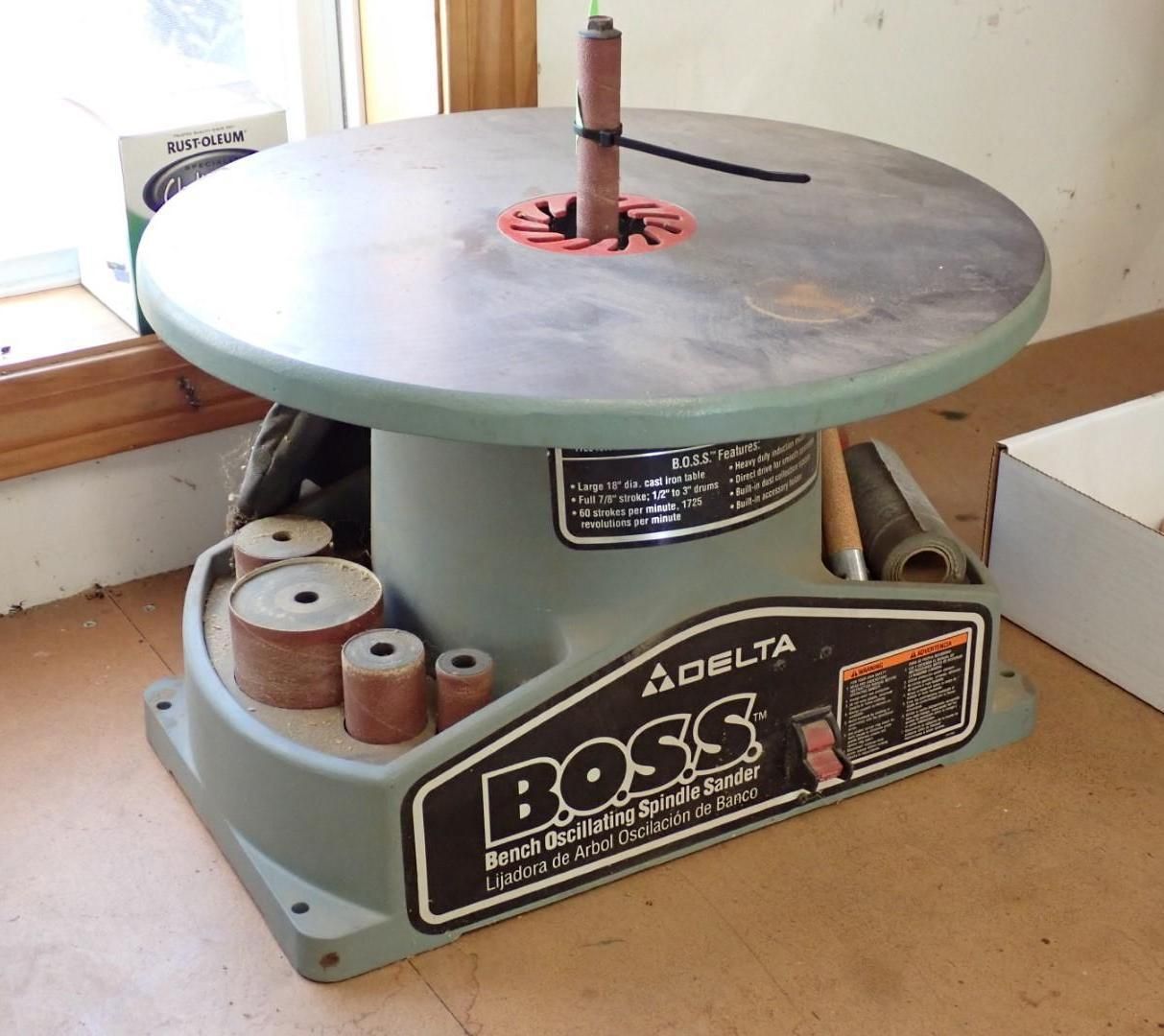 Delta BOSS oscillating spindle sander, gray and metal, on a workbench. Various sanding drums visible.