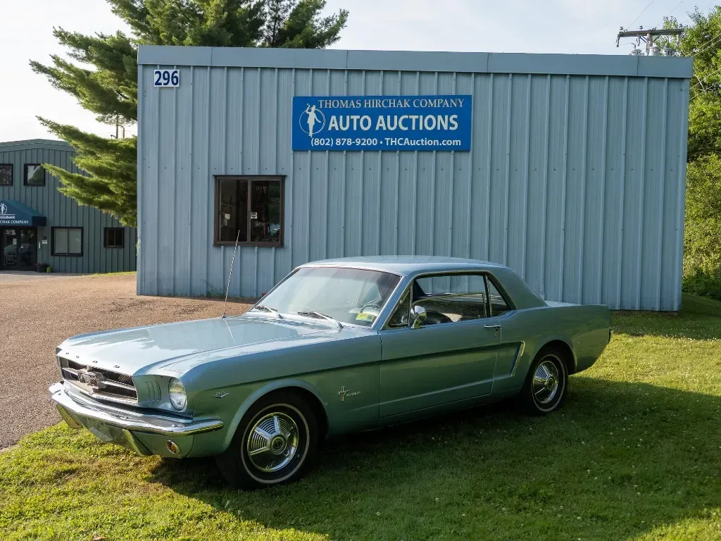 Light blue 1960s Ford Mustang parked in front of an auto auction building with a blue sign.