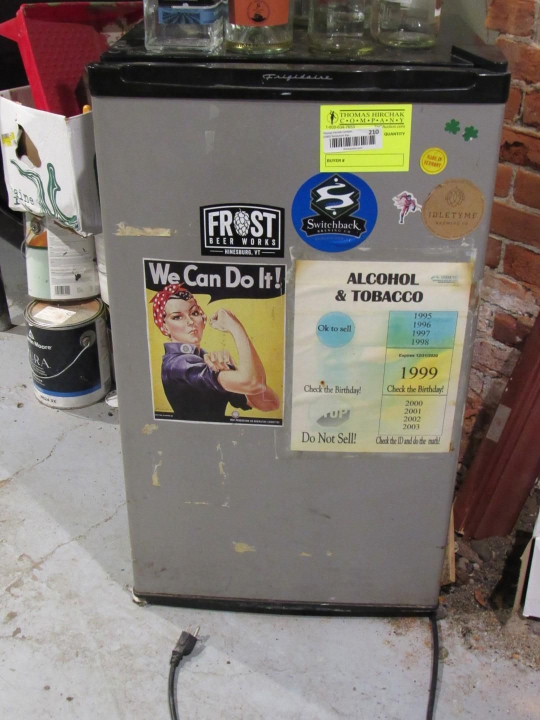 Small refrigerator with stickers, power cord, and bottles on top, in a brick-walled room.