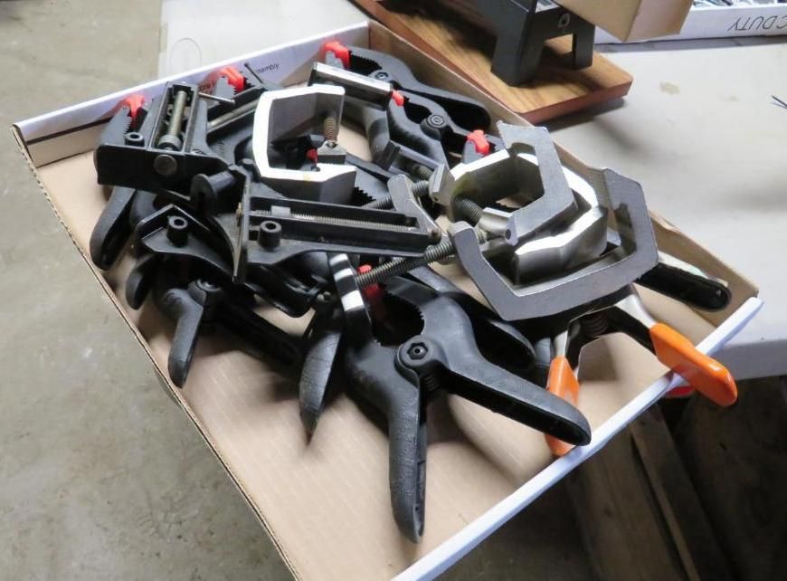 Black and orange clamps piled in a cardboard box on a workbench.