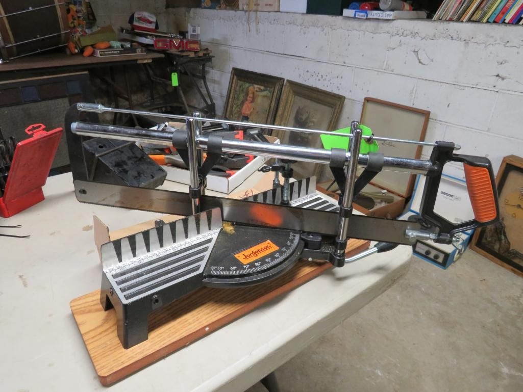 Miter saw on a wood base, on a white table. Saw blade visible, surrounded by angled guides.