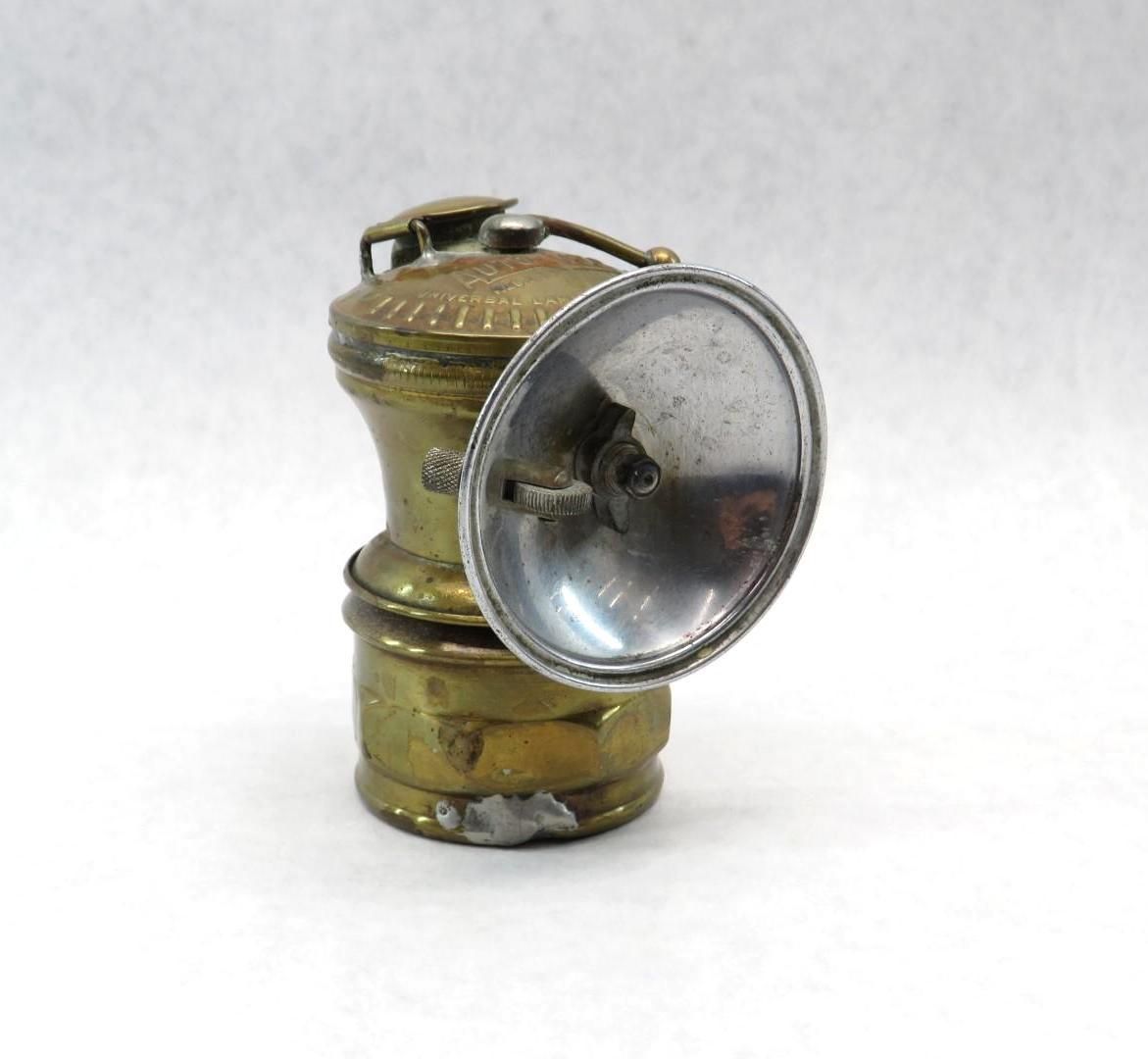 Antique brass and silver miner's lamp with a reflector, handle, and a cylindrical body, on a white surface.