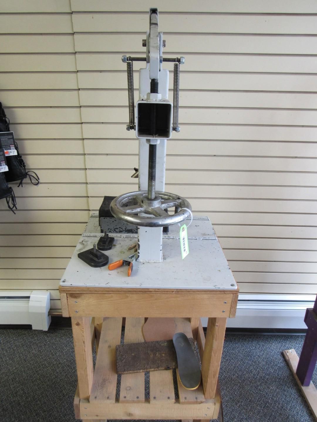 Shoe repair machine on a wooden table. The machine is grey, table is wood, and the background is a beige wall.
