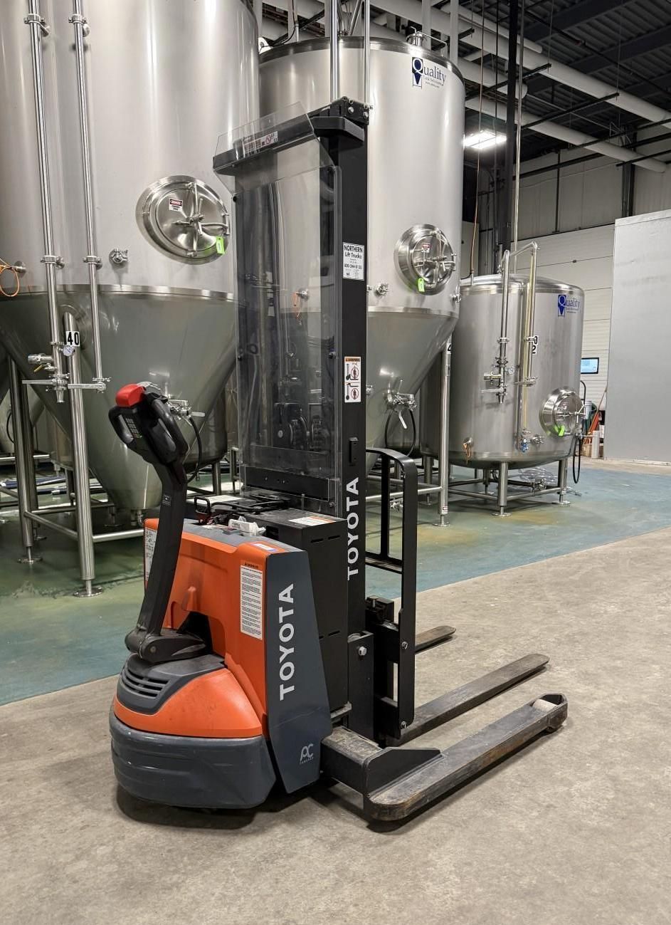 Orange and black Toyota stand-up forklift in a brewery with stainless steel tanks in the background.