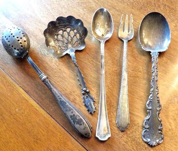 Five ornate silver spoons on a wooden surface, including a tea strainer and a serving fork.