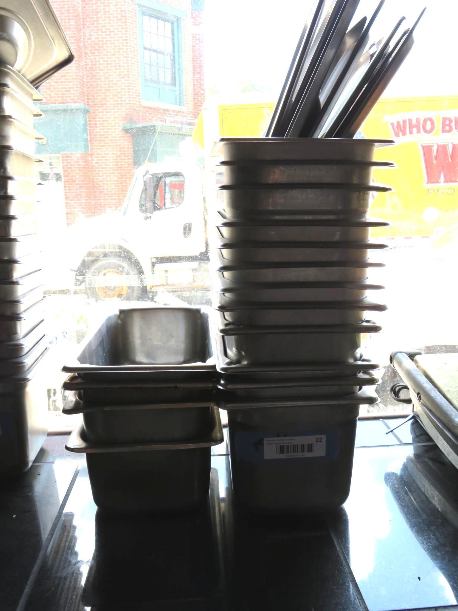 Stacks of stainless steel food pans on a counter near a window. Some are empty, while others hold utensils.