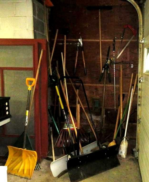 Tools leaning against a garage wall: rakes, shovels, brooms, shears, and other garden tools.