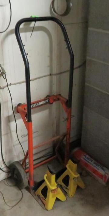 Hand truck and yellow jack stands against a wall in a basement or garage setting.