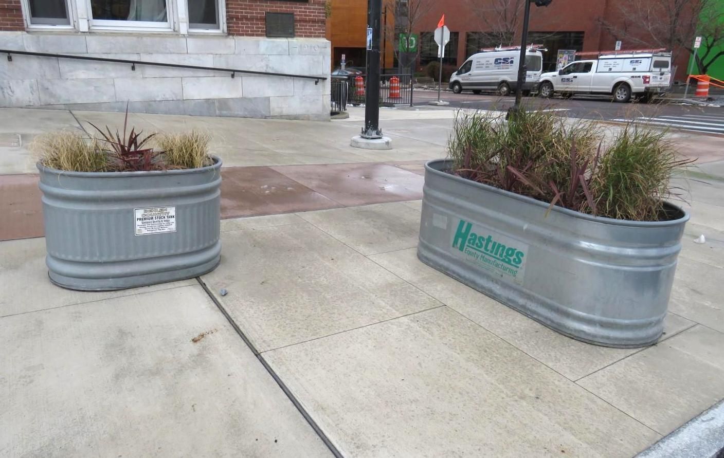 Two galvanized metal planters with plants on a sidewalk.