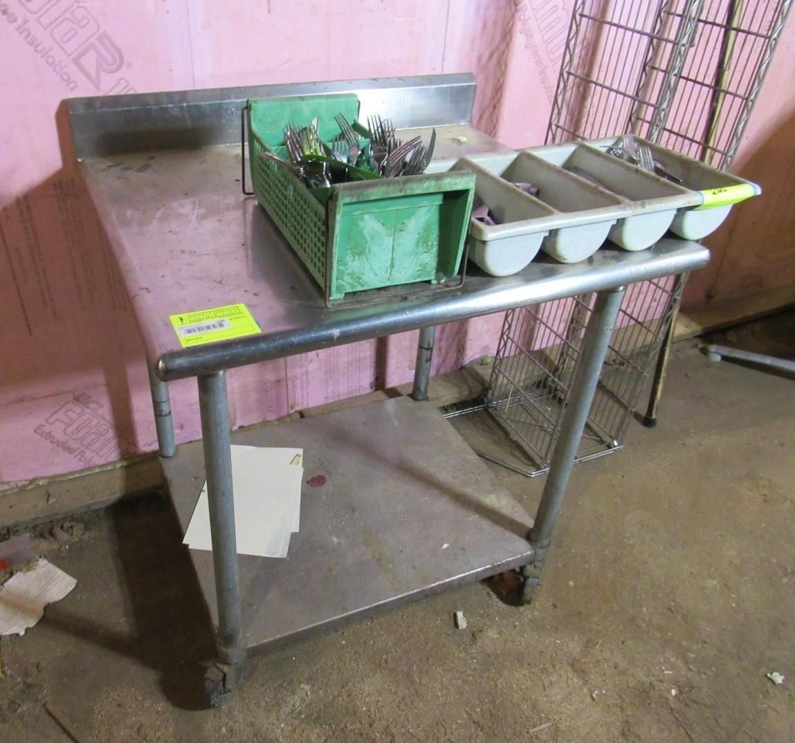Stainless steel prep table with utensils and trays on a lower shelf.