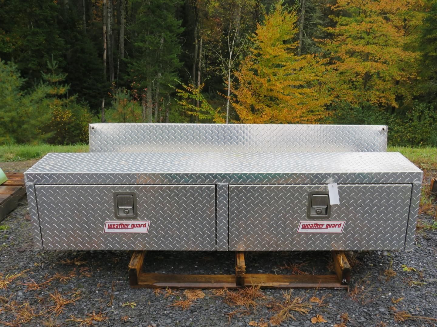 Diamond-plate metal truck bed toolbox with two drawers and a raised back section, sitting outdoors.