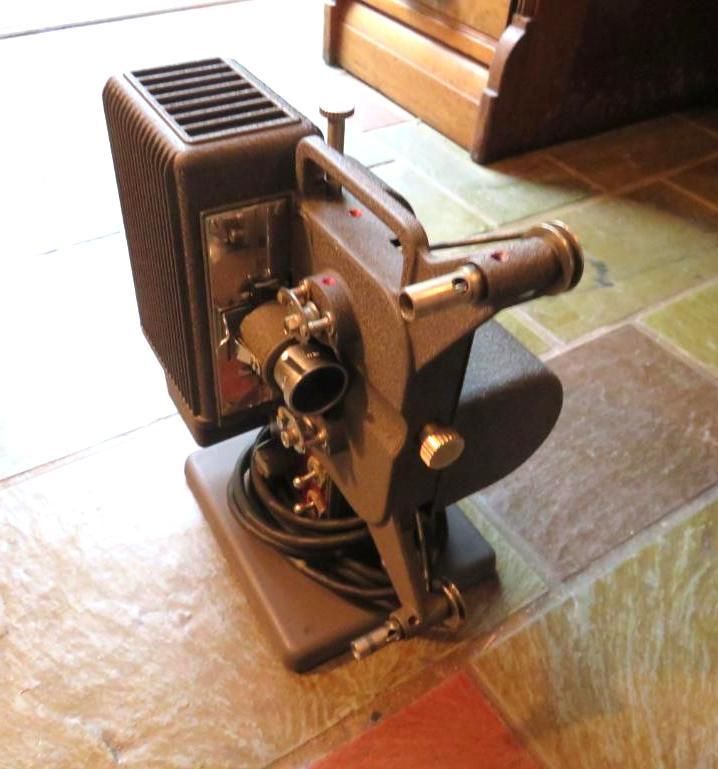 A vintage film projector, gray metal, on a tiled floor. Black cord, lens and handle visible.