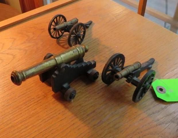 Three miniature toy cannons on a wooden table, two are black and one is brass.