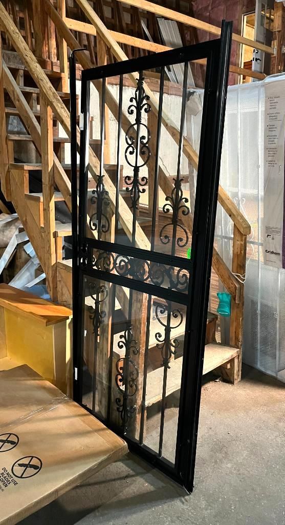 Black wrought-iron security door standing open, leaning against wooden stairs in a storage area.