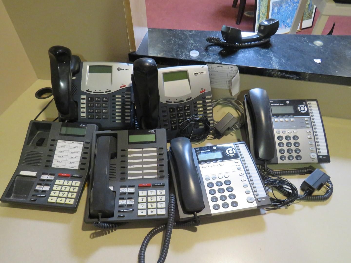 Seven different office phones of various models and colors are arranged on a counter.