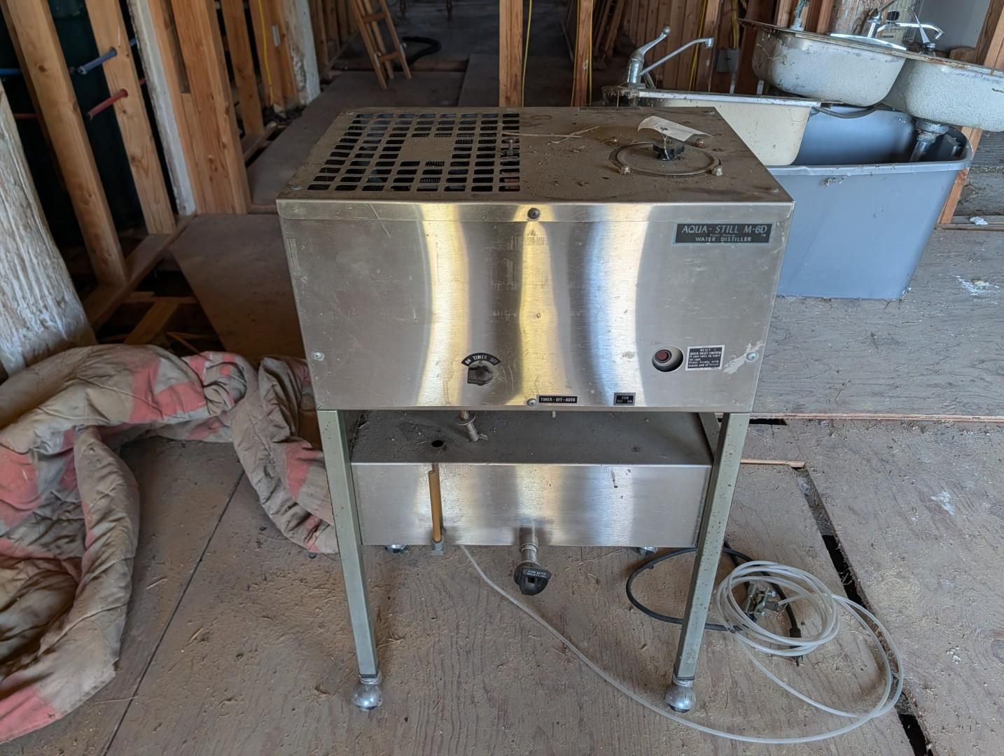 Stainless steel industrial machine on four legs in a workshop, with a coiled power cord nearby