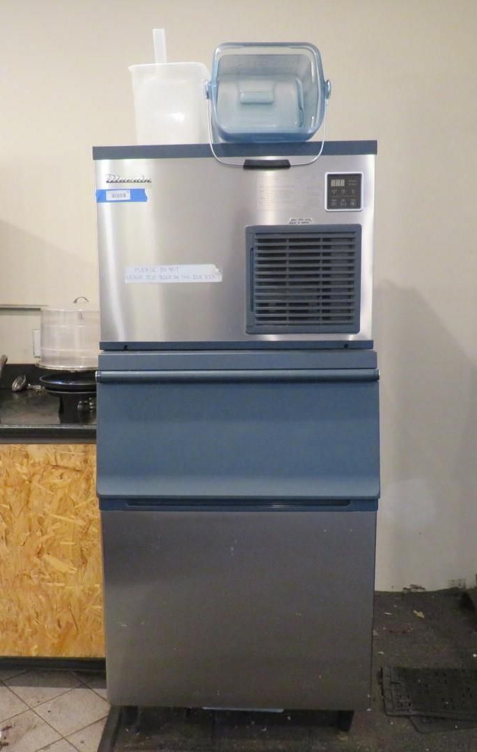 Commercial ice machine in a kitchen setting; stainless steel and blue, with containers on top.