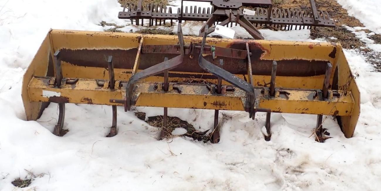 A yellow box blade attachment for a tractor sitting in the snow, featuring metal shanks and a rear grading blade.