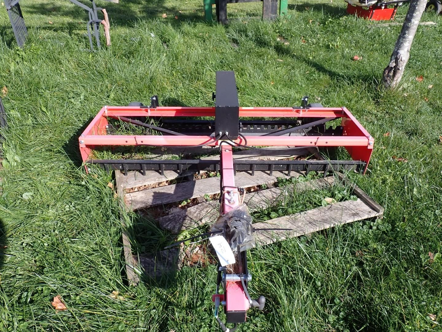 Red tow-behind mower or brush cutter resting on grass with a black hitch and wooden planks nearby