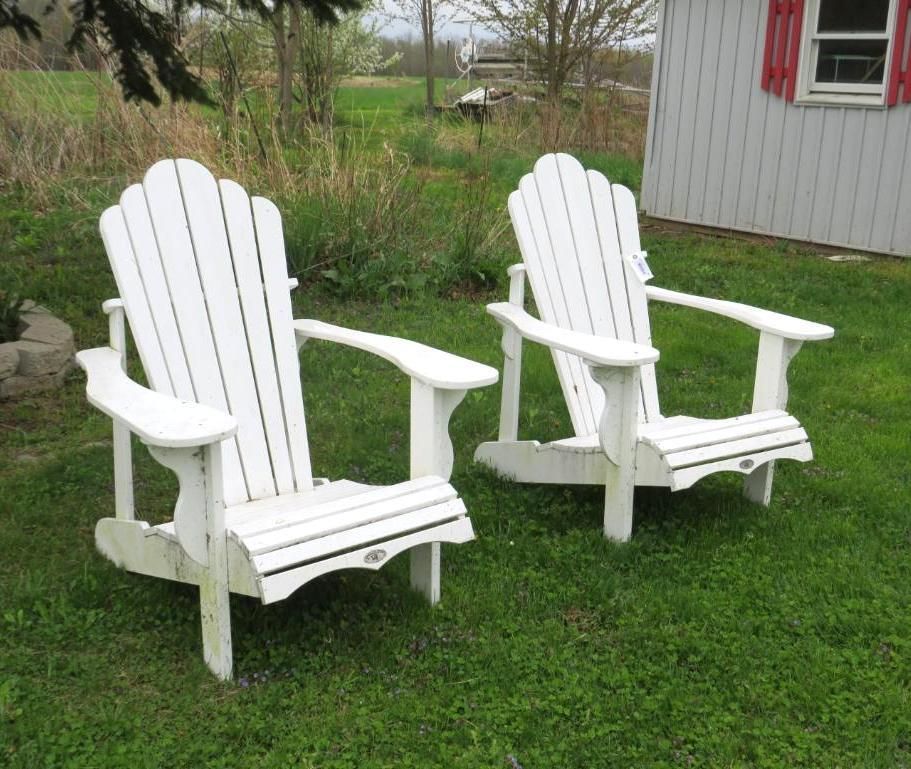 Two white Adirondack chairs on green grass in front of a white building with red shutters.