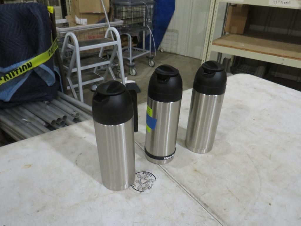 Three stainless steel travel mugs with black lids on a white table. Industrial setting.