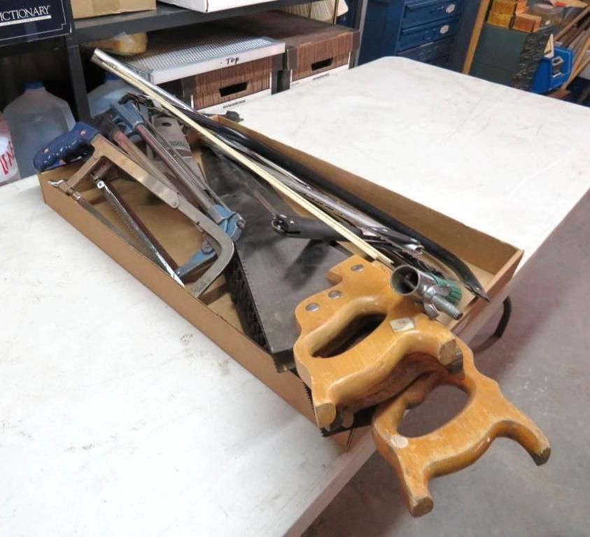Tools in a cardboard box on a light-colored table, including saws, handles, and a bow.