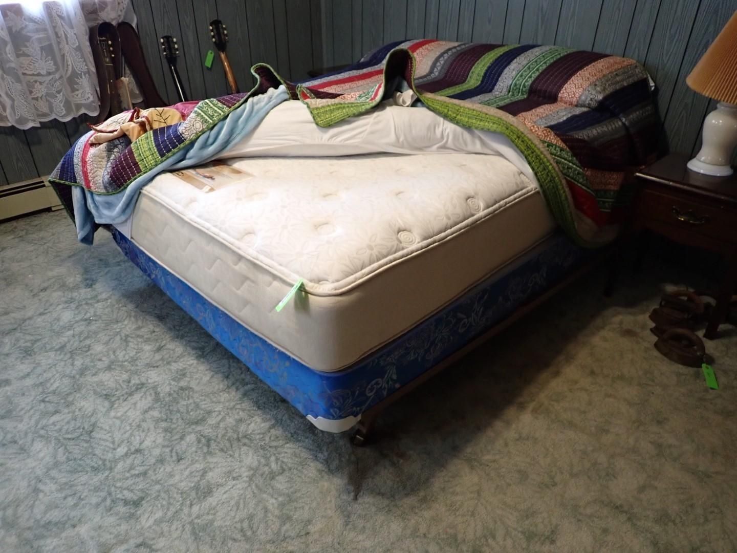 Bed with mattress and blue box spring, covered with patchwork quilts, in a room with green carpet.